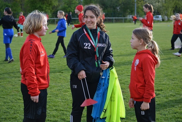 Drei M&auml;dchen stehen auf einem gr&uuml;nen Fu&szlig;ballfeld im Gespr&auml;ch; eine tr&auml;gt eine Trainerjacke und h&auml;lt Trainingsausr&uuml;stung.