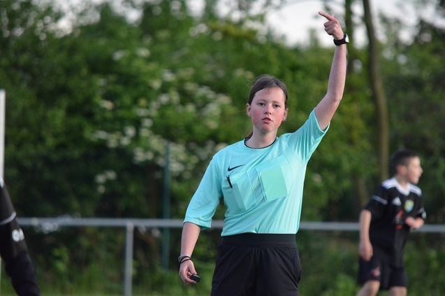 Schiedsrichterin in aquafarbenem Trikot zeigt mit dem Finger auf das Spielfeld w&auml;hrend eines Fu&szlig;ballspiels im Freien.