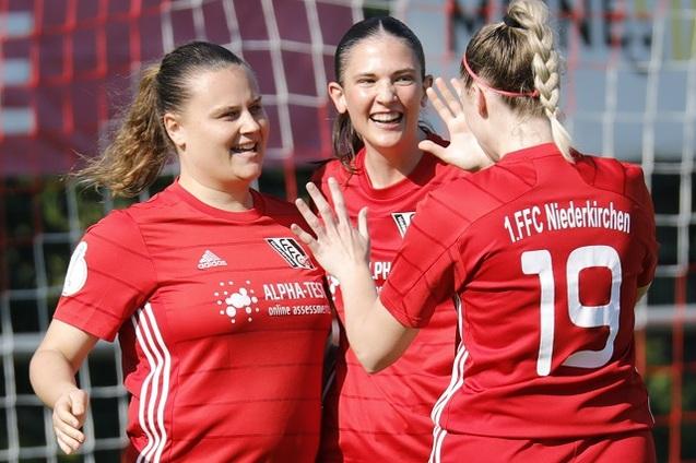 Drei Fu&szlig;ballspielerinnen in roten Trikots feiern gemeinsam auf einem Platz vor dem Tor.