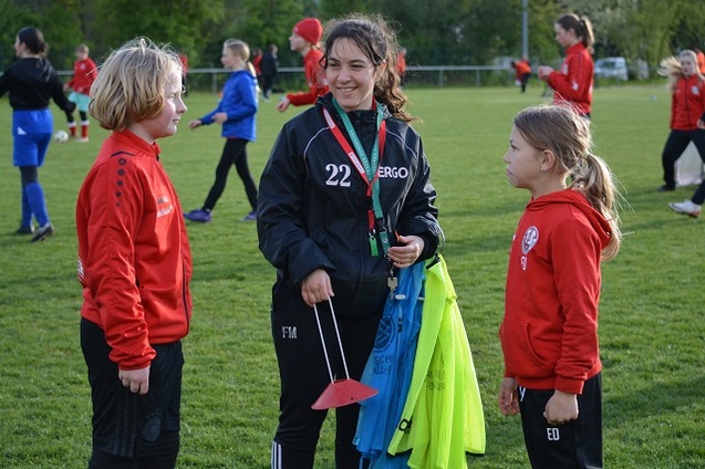 Drei M&auml;dchen stehen auf einem gr&uuml;nen Fu&szlig;ballfeld im Gespr&auml;ch; eine tr&auml;gt eine Trainerjacke und h&auml;lt Trainingsausr&uuml;stung.