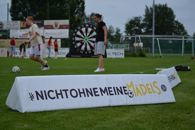 Zwei Frauen auf einem Fu&szlig;ballplatz stehen neben einem Banner mit dem Slogan &bdquo;#NICHTOHNEMEINEMADELS&ldquo;.