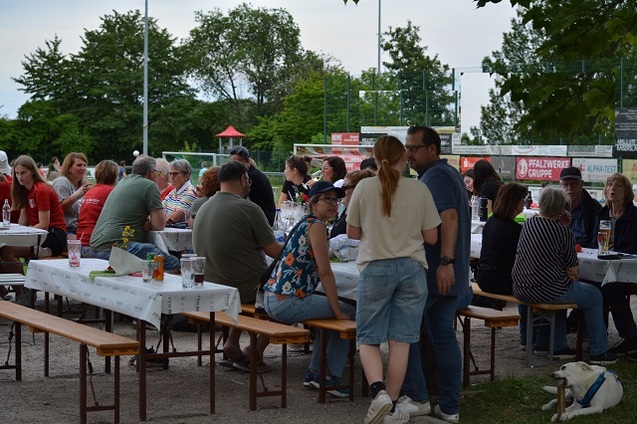Vielz&auml;hlige G&auml;ste sitzen an langen Tischen im Freien, umgeben von B&auml;umen, bei einer geselligen Veranstaltung.