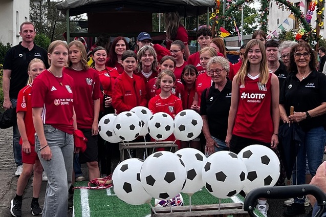 Gruppe von Menschen in roten T-Shirts mit Fu&szlig;ballmotiven posiert vor einer Dekoration mit Fu&szlig;ball-Luftballons.