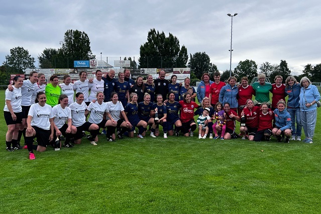 Gruppenfoto von zwei Frauenfu&szlig;ballmannschaften mit Trainern und Fans auf einer Rasenfl&auml;che, umgeben von B&auml;umen.
