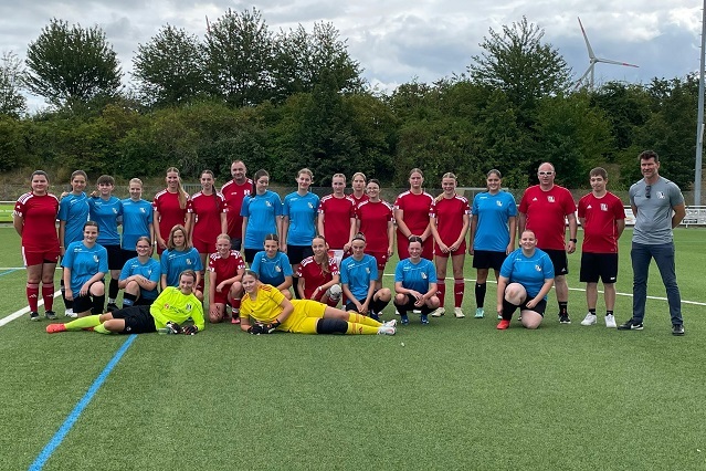 Gruppenfoto von zwei Frauenfu&szlig;ballmannschaften in rot und blau auf einem Rasenplatz, umgeben von B&auml;umen und Windr&auml;dern.
