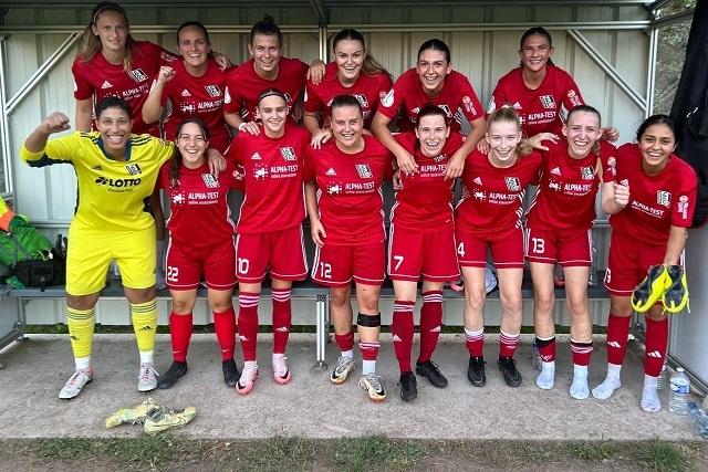 Gruppenfoto von 12 Fu&szlig;ballspielerinnen in roten Trikots, l&auml;cheln und posieren auf einer Bank. Ein Torwart steht in gelber Uniform.