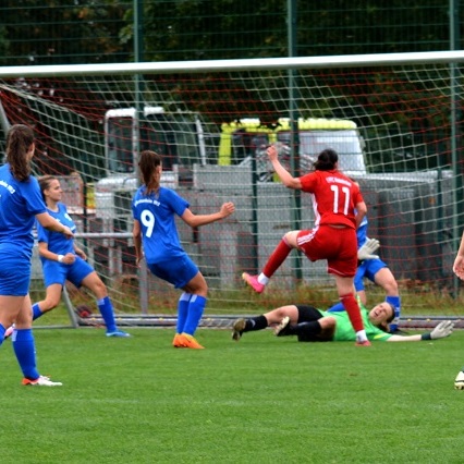 Mehrere Spielerinnen in blauen Trikots stehen um eine spielende Spielerin in rotem Trikot vor dem Tor, w&auml;hrend die Torh&uuml;terin am Boden liegt.