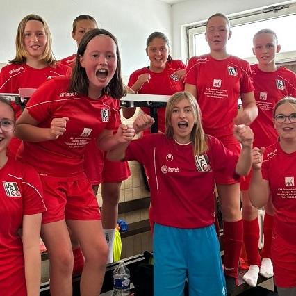 Gruppe junger Mädchen in roten Fußballtrikots posiert fröhlich in einem Umkleideraum.