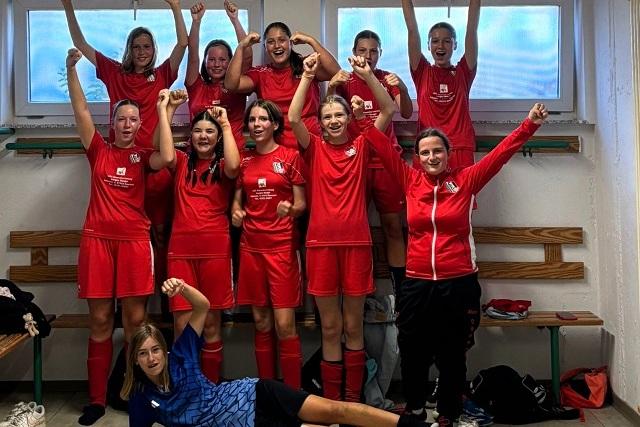 Gruppenselfie einer Mädchenfußballmannschaft in roten Trikots, die freudig die Arme heben und jubeln.