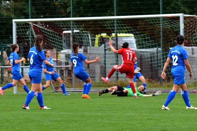 Mehrere Spielerinnen in blauen Trikots stehen um eine spielende Spielerin in rotem Trikot vor dem Tor, w&auml;hrend die Torh&uuml;terin am Boden liegt.