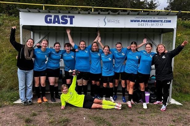 Gruppenvorstellung eines Frauenfußballteams in blauen Trikots, posierend vor einer Bank für Zuschauer.