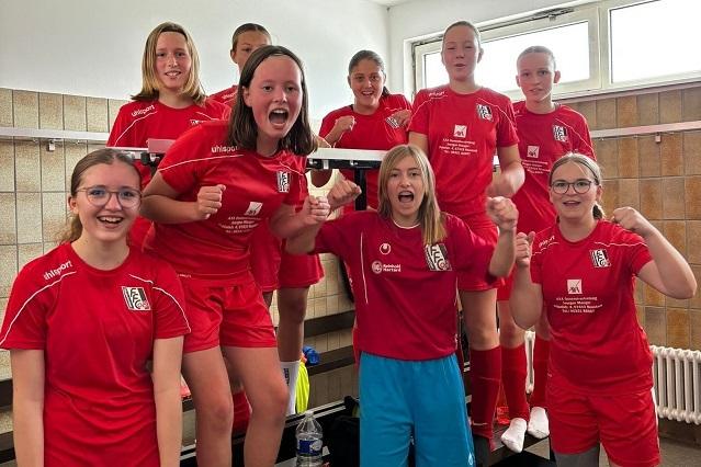 Gruppe junger Mädchen in roten Fußballtrikots posiert fröhlich in einem Umkleideraum.