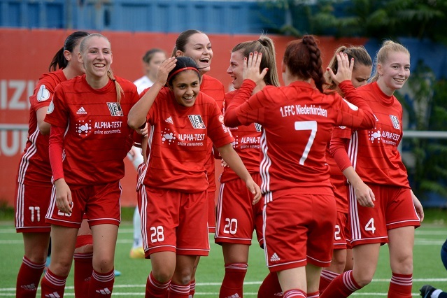 L&auml;chelnde Fu&szlig;ballspielerinnen in roten Trikots feiern gemeinsam auf einem Sportplatz.