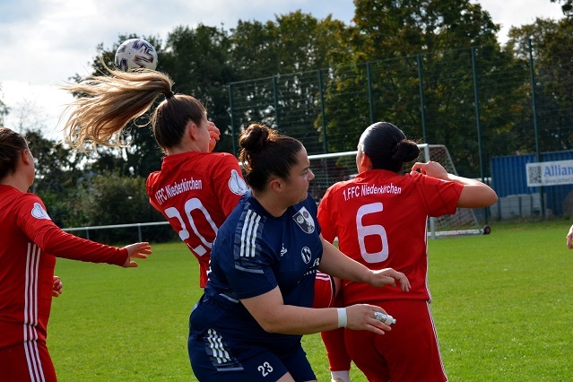 Frauenfu&szlig;ballspiel: Torh&uuml;terin in blauer Uniform rettet, w&auml;hrend Mitspielerinnen in roten Trikots um den Ball k&auml;mpfen.