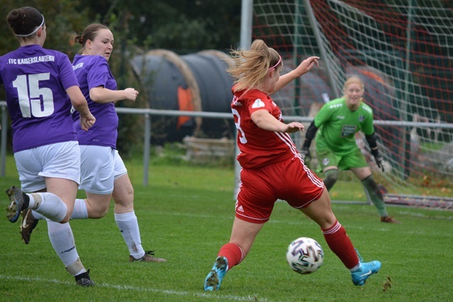 Eine Spielerin in rotem Trikot schie&szlig;t den Ball, w&auml;hrend Gegnerinnen in lila Trikots darauf reagieren. Im Hintergrund ein Torwart.