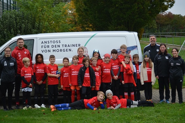 Gruppenshot von Kindern in roten Fu&szlig;balltrikots und Trainern vor einem wei&szlig;en Lieferwagen mit Motivationstext.