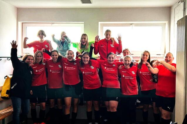 Gruppenaufnahme von Frauen in roten Fu&szlig;balltrikots, die in einer Umkleidekabine jubeln und posieren.