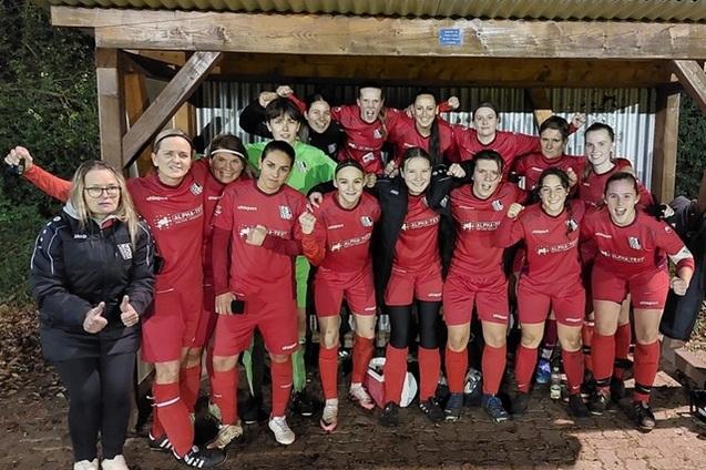 Gruppe von Fu&szlig;ballspielerinnen in roten Trikots, l&auml;chelnd und feiernd unter einem Holzunterstand.