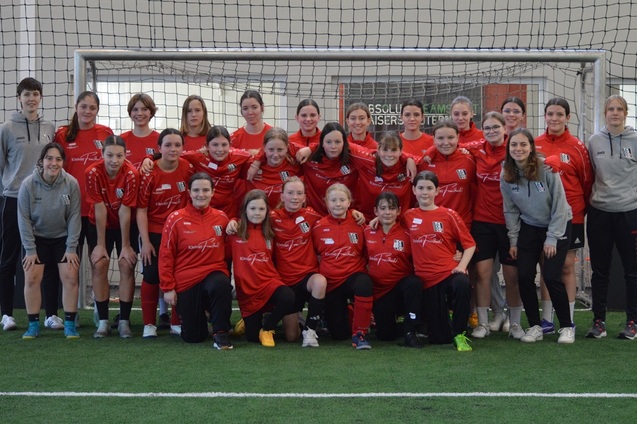 Gruppenfoto einer Frauenfu&szlig;ballmannschaft in roten Trikots, posierend auf einem Kunstrasenplatz vor dem Tor.
