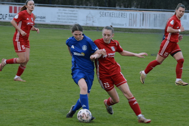 Zwei Fu&szlig;ballspielerinnen im Duell auf dem Platz, eine in Blau, die andere in Rot. Im Hintergrund l&auml;uft eine dritte Spielerin.