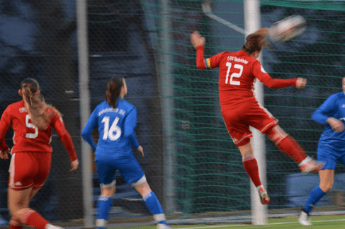 Spielszene einer Frauenfußballpartie: Spielerin in Rot springt hoch, um den Ball ins Tor zu köpfen, Torwart in Blau vorbereitet sich.