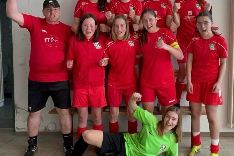 Gruppe von Mädchen in roten Fußballtrikots und Trainer, fröhlich posierend, eine Torhüterin in grün am Boden liegend.