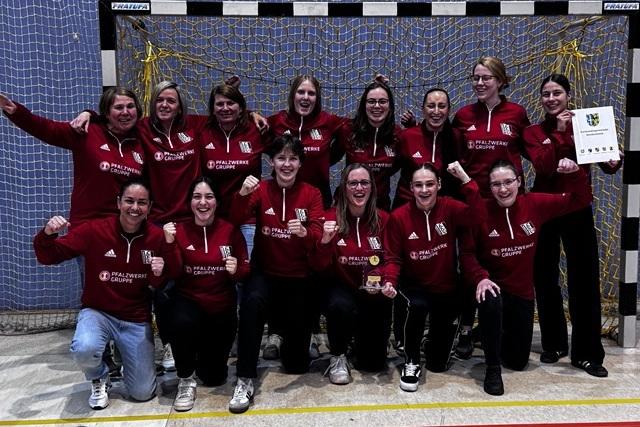 Gruppenfoto von 14 Frauen in roten Sweatshirts, jubelnd vor einem Handballtor mit Pokal und Urkunde.