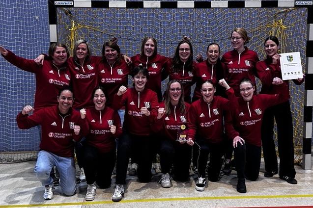 Gruppenfoto von 14 Frauen in roten Sweatshirts, jubelnd vor einem Handballtor mit Pokal und Urkunde.