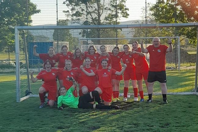 Gruppe von Fußballspielerinnen in roten Trikots posiert stolz vor einem Torfeld, ein Torwart liegt im Gras.