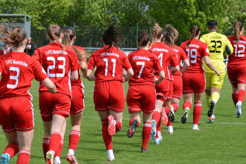 Mannschaft des 1. FFC Niederkirchen in roten Trikots, sprintend auf einem Rasenplatz w&auml;hrend eines Spiels.