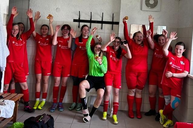 M&auml;dchenmannschaft in roten Trikots feiert fr&ouml;hlich in einer Umkleidekabine nach einem Spiel, mit Snacks in der Hand.