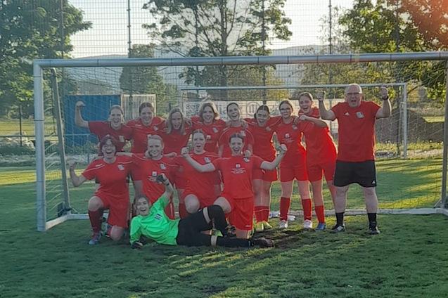 Gruppe von Fu&szlig;ballspielerinnen in roten Trikots posiert stolz vor einem Torfeld, ein Torwart liegt im Gras.