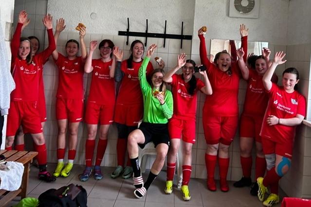 Mädchenmannschaft in roten Trikots feiert fröhlich in einer Umkleidekabine nach einem Spiel, mit Snacks in der Hand.