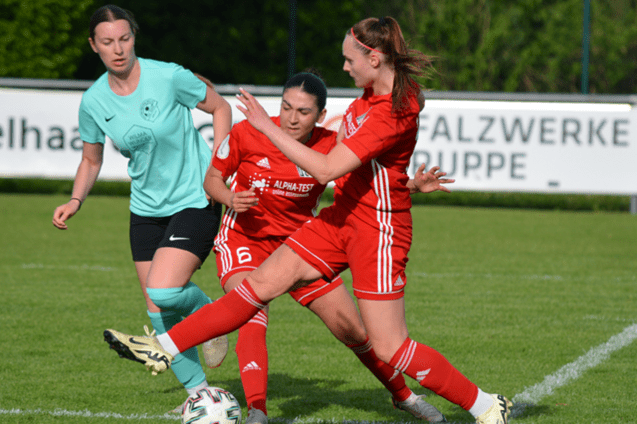 Drei Frauenfu&szlig;ballspielerinnen in roten Trikots k&auml;mpfen um den Ball auf einem gr&uuml;nen Spielfeld.