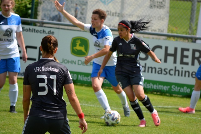 Spielszene mit zwei Frauenfu&szlig;ballmannschaften, eine Spielerin in Schwarz dribbelt den Ball, w&auml;hrend andere Spieler gestikulieren.