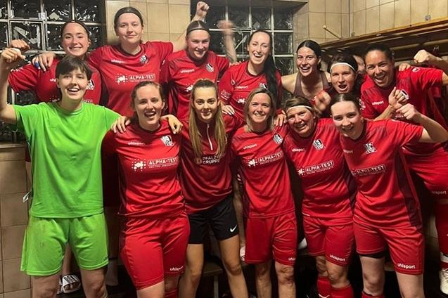 Gruppenselfie von weiblichen Fu&szlig;ballspielerinnen in roten Trikots, die fr&ouml;hlich im Umkleideraum posieren.