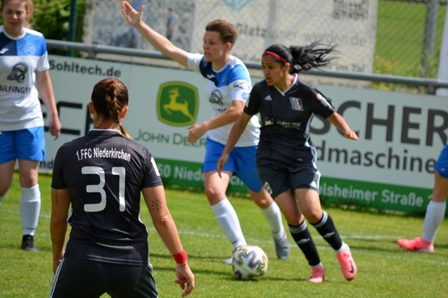 Spielszene mit zwei Frauenfu&szlig;ballmannschaften, eine Spielerin in Schwarz dribbelt den Ball, w&auml;hrend andere Spieler gestikulieren.