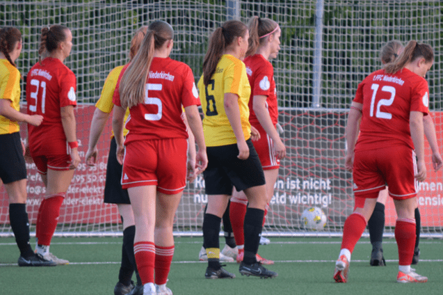 Spielszene mit Spielerinnen in roten Trikots gegen gelbe Trikots, Ergebnisanzeige 2-3, Fu&szlig;ballfeld im Hintergrund.