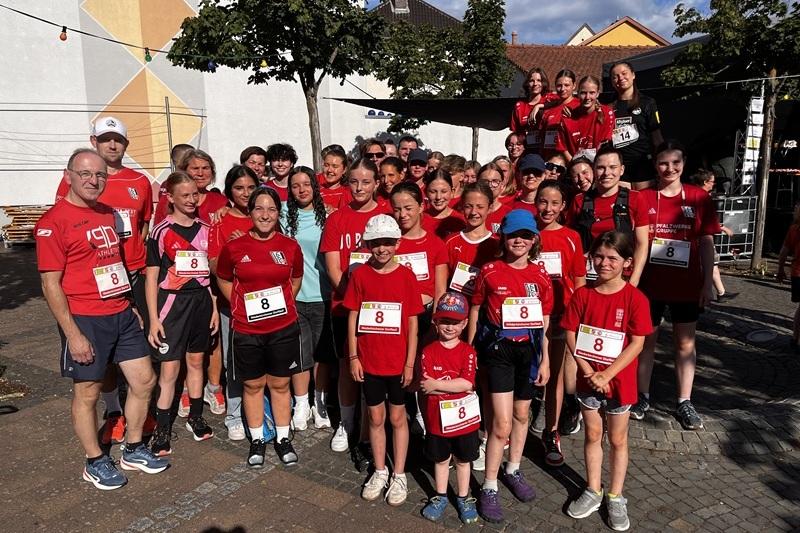 Gruppe von Läufern in roten T-Shirts mit Startnummern, auf einem Platz vor einer Wand mit buntem mural.
