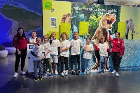 Gruppe von Kindern in T-Shirts vor einer interaktiven Klimastelle mit Dinosaurier-Illustration.