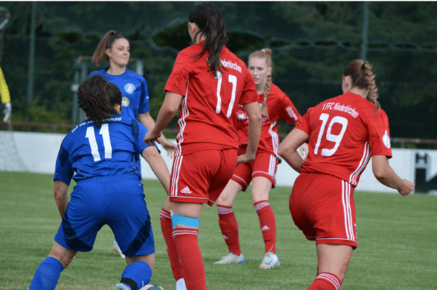 Spielszene mit Spielerinnen in roten und blauen Trikots auf einem Fu&szlig;ballfeld, Ergebnis und Torsch&uuml;tzen eingeblendet.
