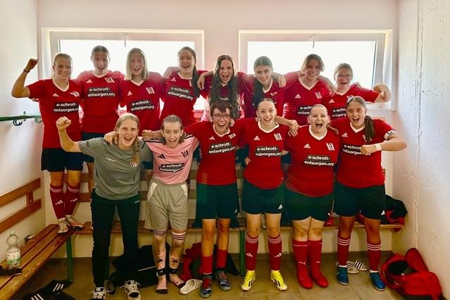 Gruppe von Fu&szlig;ballspielerinnen in roten Trikots, die jubelnd im Umkleidebereich posieren.