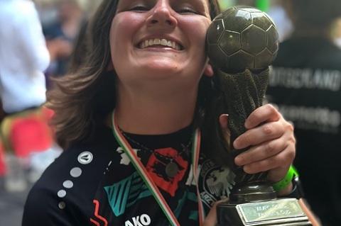 Junge Sportlerin h&auml;lt stolz einen Pokal in Form eines Fu&szlig;balls und l&auml;chelt, umgeben von festlicher Atmosph&auml;re.