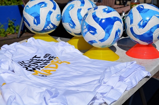 Wei&szlig;e T-Shirts mit schwarzer und gelber Schrift liegen neben mehreren Fu&szlig;ballen auf einem Tisch mit farbigen St&auml;ndern.