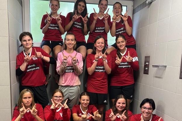 Gruppentreffen von 12 Fußballspielerinnen in roten Trikots, die in einem Umkleideraum Fröhlichkeit zeigen.