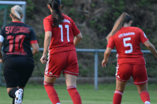 Zwei Fußballspielerinnen in roten Trikots laufen auf einem Spielfeld, während eine Spielerin in Schwarz zurückblickt.