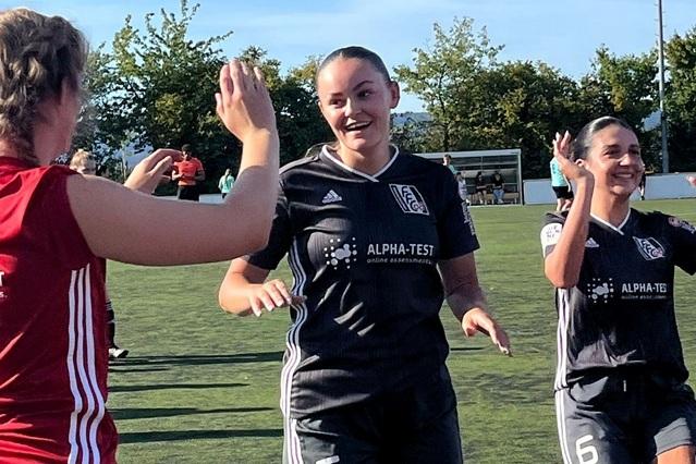 Zwei Fu&szlig;ballspielerinnen in Sportkleidung feiern einen erfolgreichen Moment auf dem Platz, umgeben von B&auml;umen und Zuschauern.