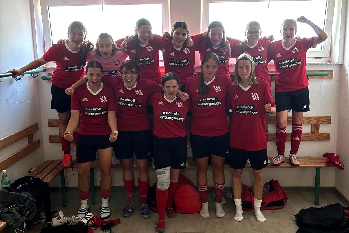 Gruppe von zwölf Fußballspielerinnen in roten Trikots, fröhlich posierend in einer Umkleidekabine.