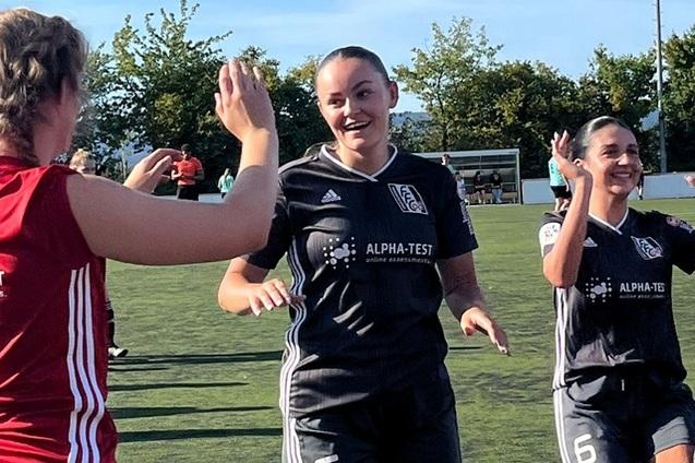 Zwei Fußballspielerinnen in Sportkleidung feiern einen erfolgreichen Moment auf dem Platz, umgeben von Bäumen und Zuschauern.