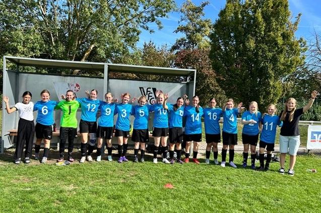 Mannschaft von 15 Fu&szlig;ballspielerinnen in blauen Trikots posiert l&auml;chelnd auf einem Sportplatz unter blauem Himmel.